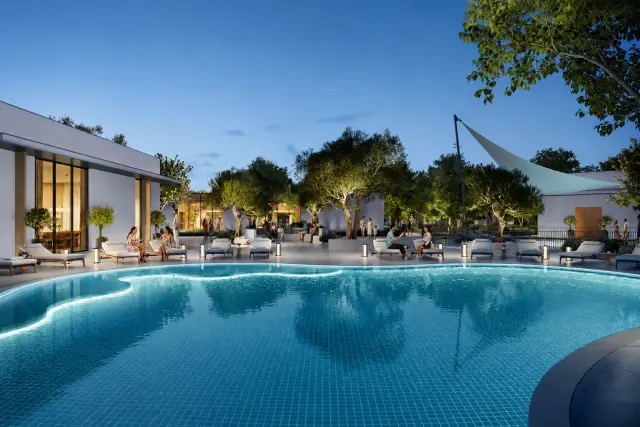 Illuminated curvilinear swimming pool with white lounge chairs and lush trees at Alva, The Valley by Emaar at twilight